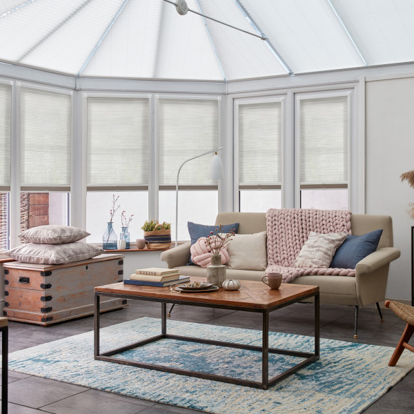 White pleated cellular blinds on conservatory windows and roof, closed and softly diffusing daylight; cozy sunroom living area with beige sofa, chunky pink throw, wooden coffee table on blue rug.