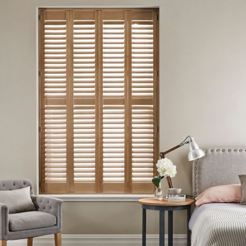 Wooden plantation shutters covering tall recessed window, fixed and tilted to let soft light through; bedroom setting with upholstered bed, bedside table lamp, flowers, armchair in neutral tones.