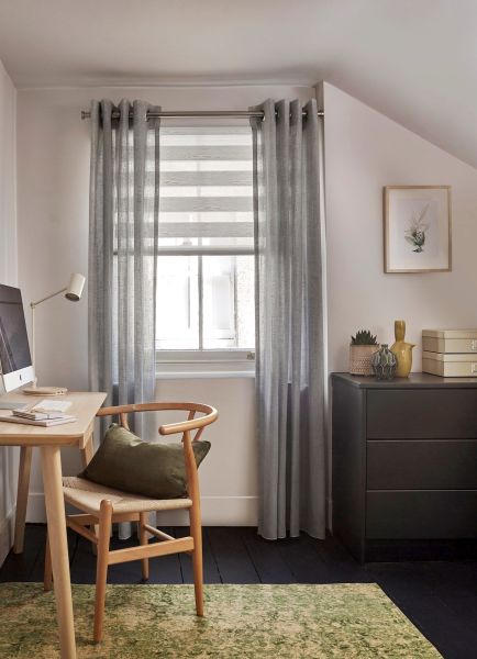 Sheer grey grommet curtains frame a window with a partially lowered striped roller blind, filtering daylight into compact home office with a wooden desk, chair, dark dresser and green rug.