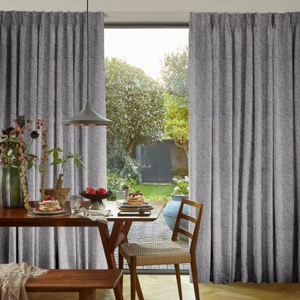 Full-length gray geometric pleated curtains, hung from a rod, pulled aside at center to reveal a sunlit backyard; wooden dining table with pancakes, chairs and pendant lamp occupy the foreground.