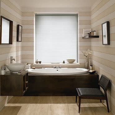 White horizontal blinds, fully lowered and closed, filtering soft daylight through slats; covering a central bathroom window above a dark-wood bathtub. Neutral striped walls, vessel sink, chair, shelf with toiletries.