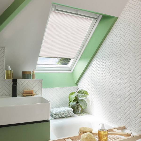 Pleated skylight blind in pale pink, mounted inside a roof window and lowered to filter daylight; sloped-ceiling bathroom with mint-green trim, white herringbone tiles, sink, bathtub and potted plant.