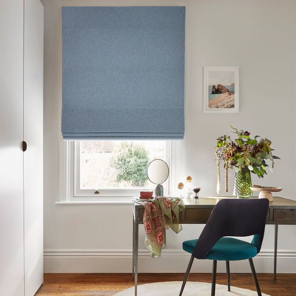 Light-blue Roman blind, lowered in soft folds, filtering daylight. Above a white sash window over a wooden desk with chair, small mirror and vase of flowers in a neutral-painted room.