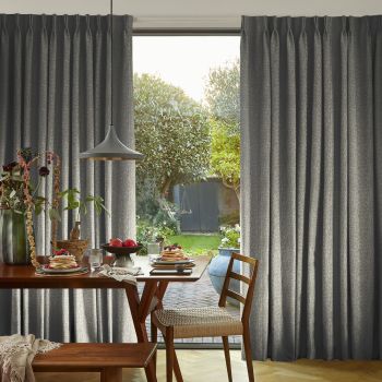 Curtains: floor-to-ceiling grey pleated drapes with textured fabric, framing a window; Action: parted centrally, softly filtering daylight; Context: dining table with pancakes, plants and pendant lamp, sunlit garden beyond.