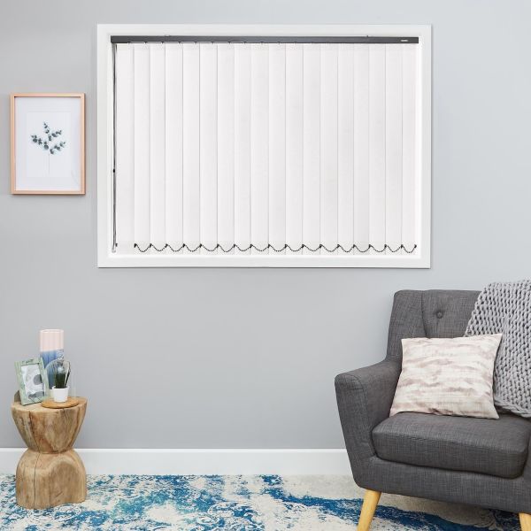 White vertical blinds covering a window, closed with bead chain along bottom, softly filtering light; mounted on a light-gray wall above a gray armchair, wooden side table and blue rug.