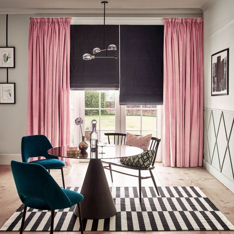 Pink pleated floor-length curtains frame a large window with two black Roman blinds partially lowered, filtering daylight; dining area with round glass table, teal chairs, striped rug and garden view.