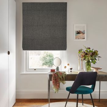 Dark grey Roman shade, fully lowered with subtle folds at the bottom, filtering daylight through a white sash window; above a wooden desk with teal chair, vase of flowers and a small mirror.