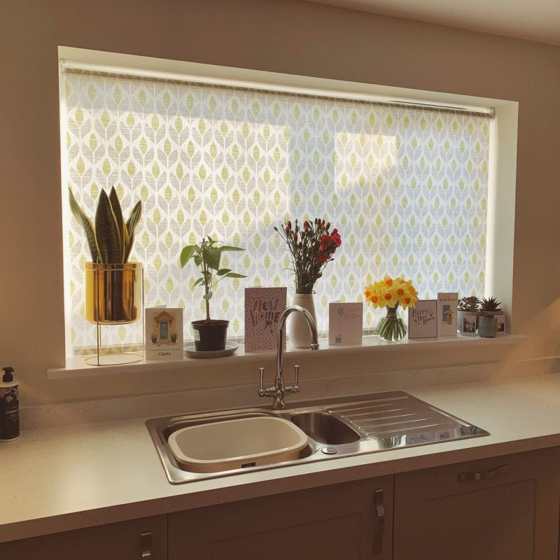 Patterned roller blind, lowered and semi‑translucent, filtering daylight over a kitchen sink; windowsill holds potted plants, vases of flowers, and several greeting cards.
Clarity
New Home
Happy New Home
COWSHED