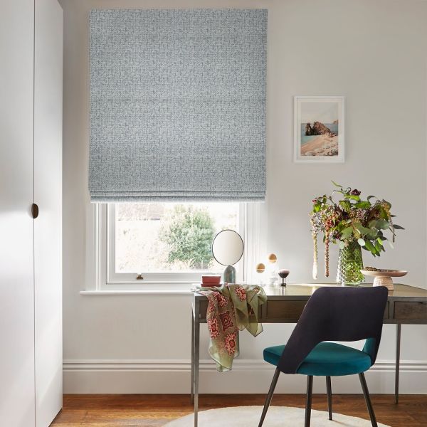 Roman blind, pale blue textured fabric, partially lowered, filtering soft daylight into a small home office with wooden desk, teal chair, vase of flowers and framed coastal print.