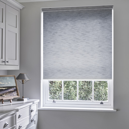 Grey textured roller blind lowered over upper two-thirds of a white sash window, partially closed and filtering daylight; pale-gray cabinetry, countertop with lamp and painting, hedge visible below.