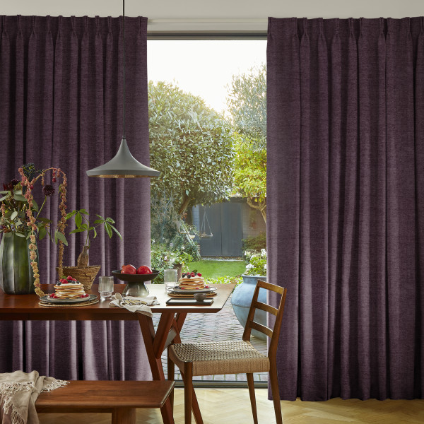 Heavy pleated purple curtains, drawn partially aside to reveal a glass sliding door; curtains frame and filter daylight into a dining area with wooden table, pendant lamp, and garden beyond.
