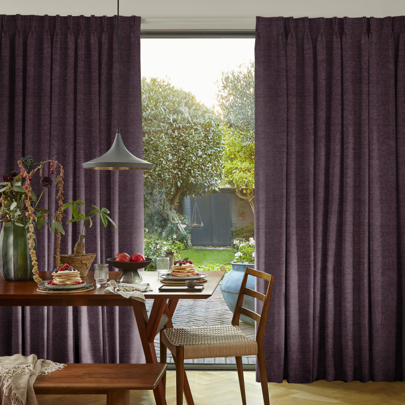 Heavy pleated purple curtains, drawn partially aside to reveal a glass sliding door; curtains frame and filter daylight into a dining area with wooden table, pendant lamp, and garden beyond.