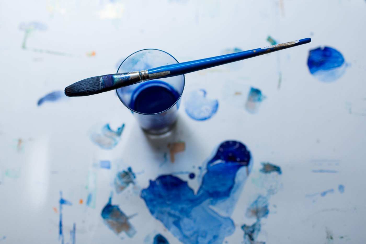 a blue paint brush and a glass of paint water on a white surface with blue paint on it.