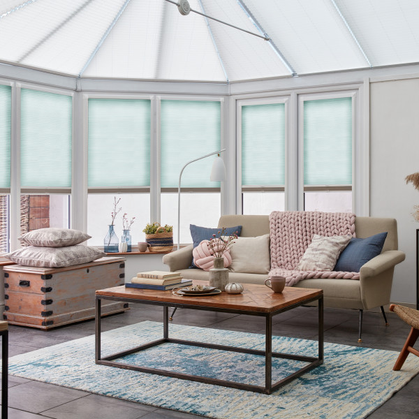 Teal pleated honeycomb blinds covering tall bay/conservatory windows and roof panels, lowered and softly filtering daylight; conservatory living room with beige sofa, pink chunky throw, wooden coffee table and rug.