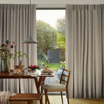 Floor-length, pinch-pleat grey curtains, drawn open at the center and hanging straight, framing a sunlit backyard; a wooden dining table set for breakfast with pancakes and a pendant light in foreground.
