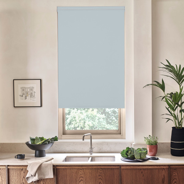 Light blue roller blind lowered over a window above a kitchen sink, blocking light; stainless faucet centered on a marble countertop with wooden cabinets, potted plants and bowls nearby.