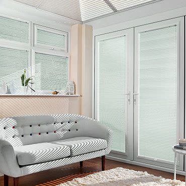 White pleated blinds on French doors and upper windows, fully lowered and producing narrow striped shadows—filtering sunlight into a bright living room with a pale tufted sofa, hardwood floor, and rug.