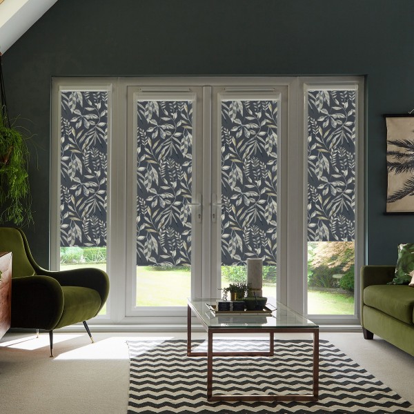 Patterned roller blinds cover French glass doors, partially lowered, in a modern living room with green armchairs, a glass coffee table on a chevron rug, and a garden visible outside.