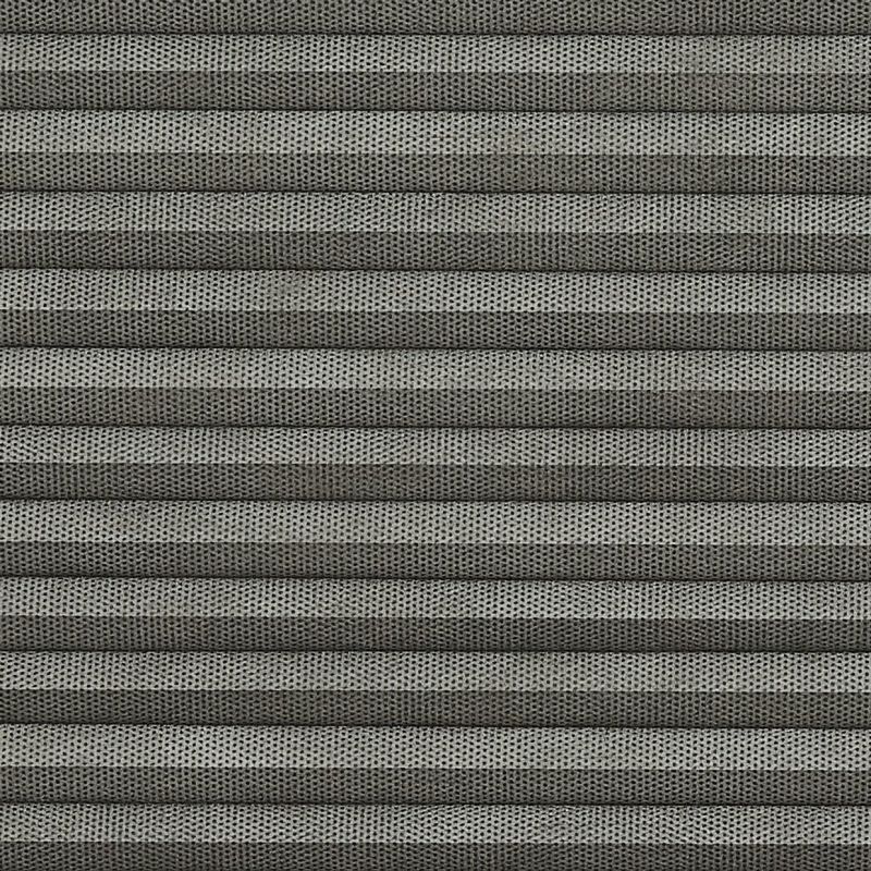 Grey pleated cellular blind, fully closed, narrow horizontal folds with perforated textured fabric; filtering light and covering a window in a softly lit interior, no visible view.