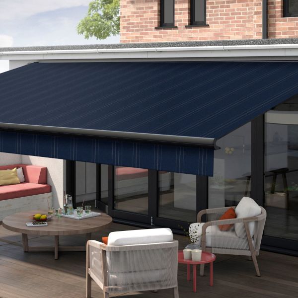 A navy-blue fabric retractable awning with subtle diagonal stripes and matching valance, extended over glass patio doors, shading a sunlit outdoor seating area with table, chairs, brick wall and tree.