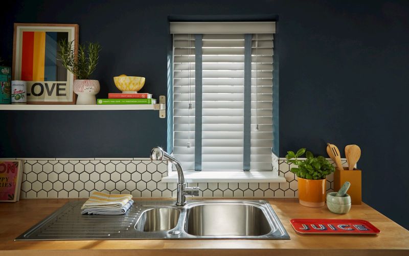 White horizontal Venetian blinds with blue fabric tapes, lowered and partially closed, filtering daylight through slats; kitchen sink with dark blue walls, wooden countertop, hex-tile backsplash, plants and utensils.

Text: LOVE
Text: JUICE
Text: ROSA'S THAI CAFE THE VEGETARIAN COOKBOOK
Text: CRAFT BEER