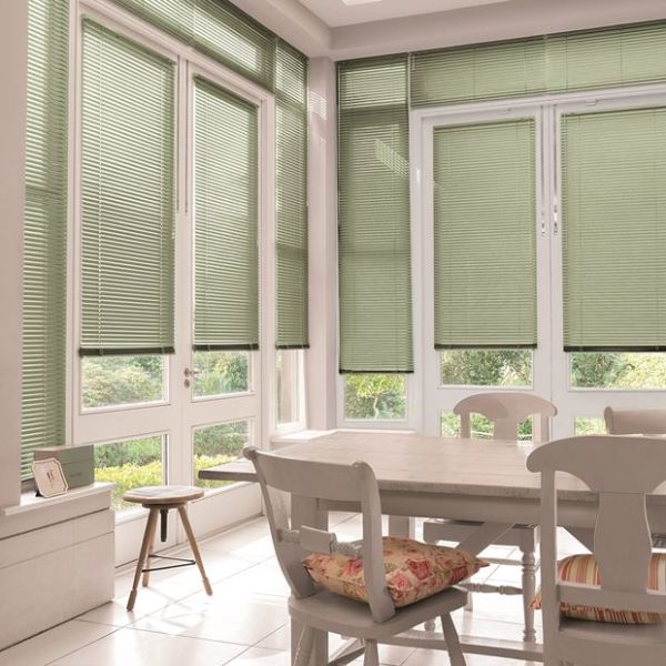 Green Venetian blinds on tall windows and patio doors, lowered and filtering daylight while leaving lower panes visible; bright sunroom dining area with white table, chairs, cushions, garden view.