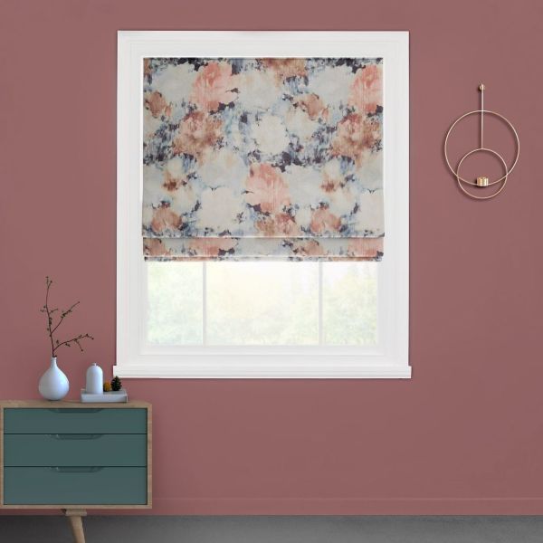 Floral-patterned Roman shade, partially lowered with visible soft folds, diffusing daylight; installed in a white-trimmed window set in a mauve wall, above a teal dresser and gold circular wall sconce.