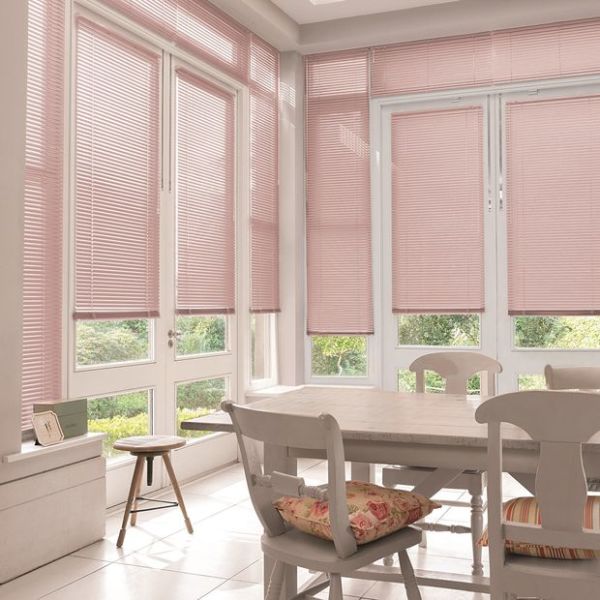 Pink horizontal Venetian blinds lowered over tall paneled windows and French doors, slats mostly closed, filtering daylight into a bright dining nook with wooden table, chairs and garden glimpses.