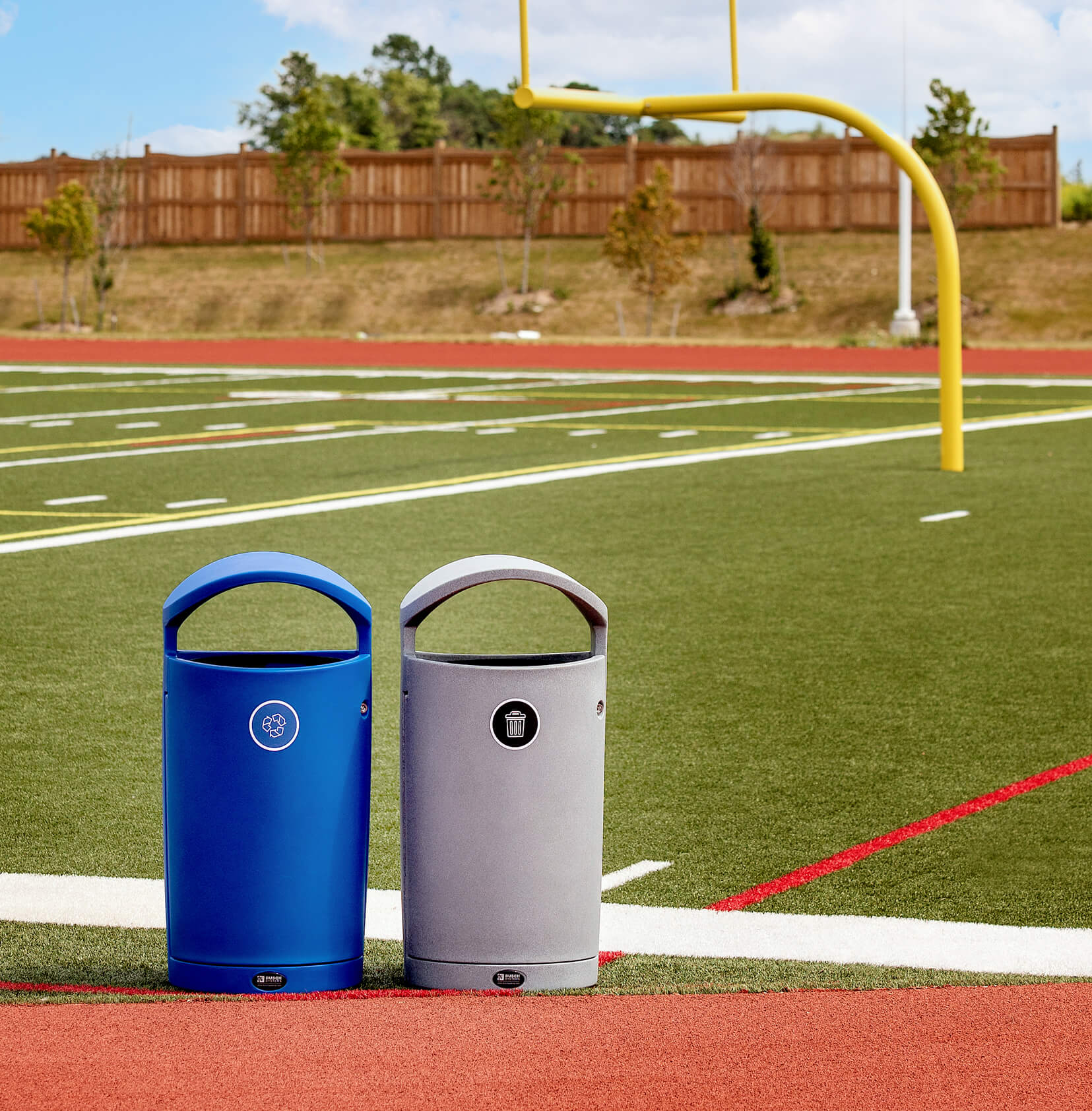 blue recycling and grey waste bins outdoors on the side of a football field