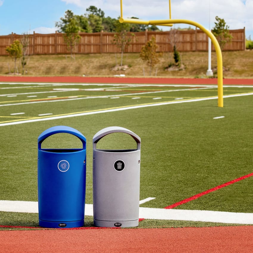 blue recycling and grey waste bins outdoors on the side of a football field