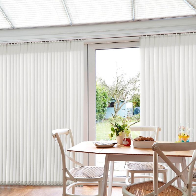 White vertical blinds, floor-to-ceiling, parted at center; filtering daylight and partially open to a glass sliding door; sunlit dining area with wooden table, chairs, and garden view.