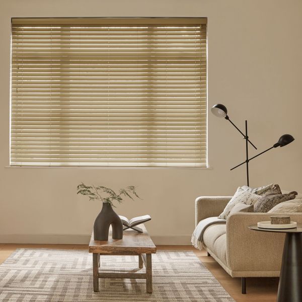 Beige horizontal venetian blinds covering the window, slats mostly closed and filtering soft daylight; above a neutral living room with beige sofa, wooden coffee table and a black floor lamp.