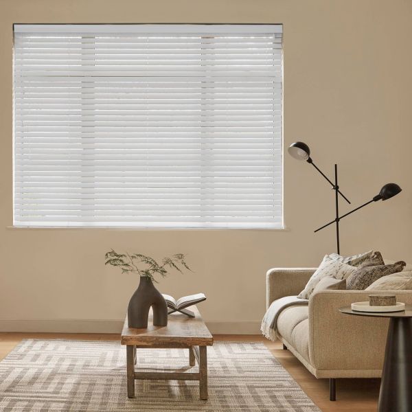 White horizontal blinds covering a rectangular window, closed and filtering soft daylight, in a beige living room with a sofa, wooden coffee table holding a vase and open book.
