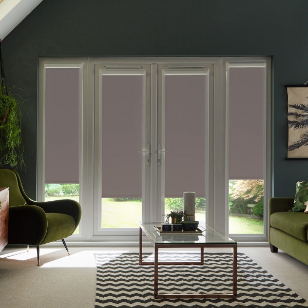 French glass doors with four mauve roller blinds lowered halfway, framing a sunlit garden; glass coffee table and zigzag rug sit between two green armchairs in a contemporary living room.