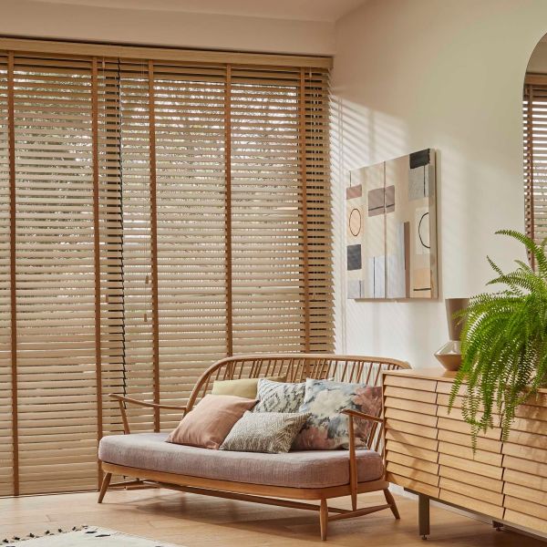 Wooden horizontal blinds covering a full-height window, slats tilted to filter sunlight — casting striped shadows across a living room with a rattan loveseat, cushions, wooden sideboard and fern.