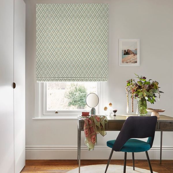 Roman blind with pale green-and-cream geometric pattern, lowered over the window, filtering soft daylight into a home office with desk, teal chair, vase of foliage, mirror and coastal print.