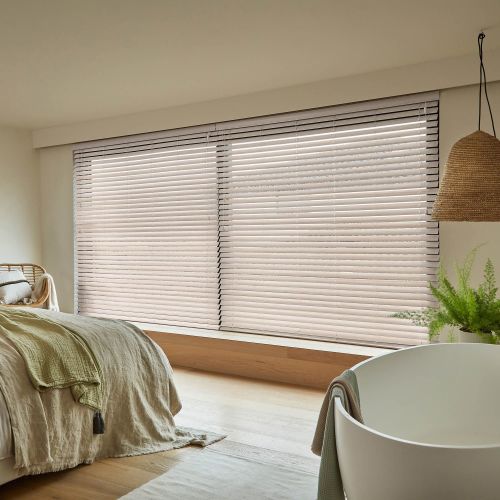 Horizontal Venetian blinds covering a wide bay window, slats closed and softly diffusing daylight; neutral, minimal bedroom with linen-covered bed, wooden floor, woven pendant light and a potted fern.