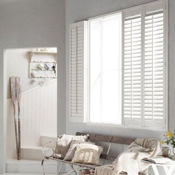 White louvered plantation shutters (three-panel): left panel folded open, right panels closed, partially filtering bright daylight over a bench with neutral cushions and throws in a light, coastal-style sitting area.