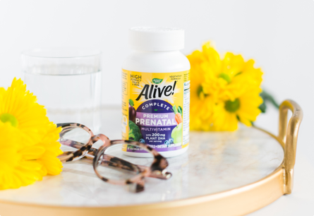 A bottle of Alive Complete Premium Prenatal Multivitamin on a marble tray with a pair of glasses and bright yellow flowers.