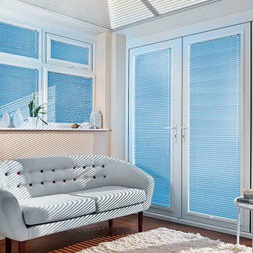 White horizontal blinds on bay windows and French doors, closed but filtering sunlight; striped light falls across tufted white loveseat in bright living room with wooden floor and rug.