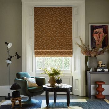 Brown patterned Roman blind (diamond motif) lowered over a white-framed sash window, filtering daylight; set in a cozy living room with green armchair, low carved table, lamp, plants, framed portrait.