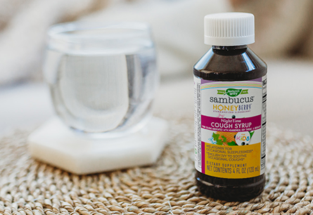 A bottle of Nature's Way Sambucus Kids HoneyBerry Night Syrup is on a woven rattan next to a glass of water.