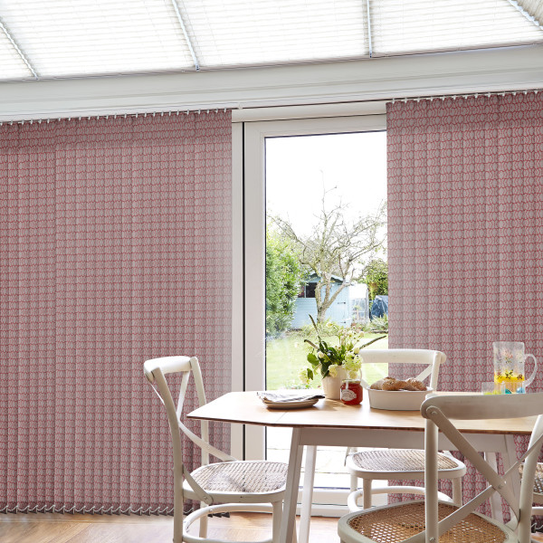 Patterned pink vertical blinds span a patio door, partially drawn to either side, filtering daylight; dining table with chairs, flowers, bread and jug sits inside, bright garden visible outside.