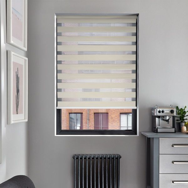 A beige zebra roller blind covers a recessed window, alternating opaque and sheer horizontal bands partially filtering light; set in a gray kitchen with espresso machine, radiator and framed art.