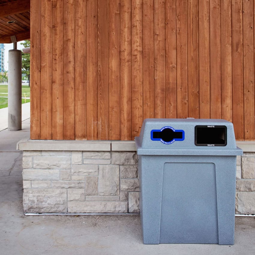 double mixed recycling and trash container with a grey body and hinged lid in front of a wooden building