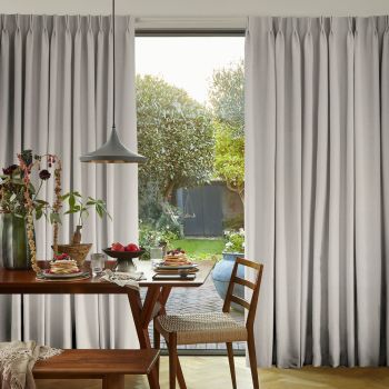 Full-length grey pleated curtains pulled aside to frame a sliding glass door, filtering soft daylight; wooden dining table set for breakfast with pancakes, a pendant lamp, and a green garden beyond.