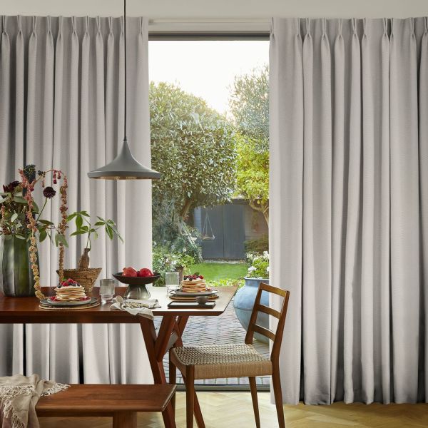 Full-length grey pleated curtains pulled aside to frame a sliding glass door, filtering soft daylight; wooden dining table set for breakfast with pancakes, a pendant lamp, and a green garden beyond.