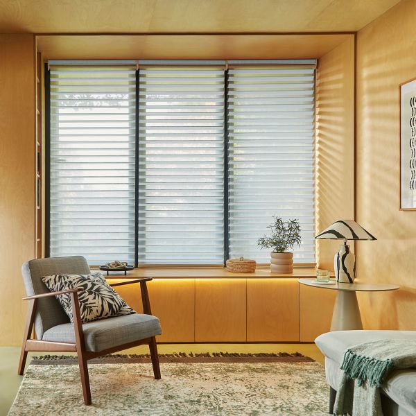 Layered horizontal sheer-and-opaque roller shades covering three tall window panels, partially closed and filtering soft daylight; set in a warm plywood-walled sitting nook with armchair, side table, lamp, and plant.
