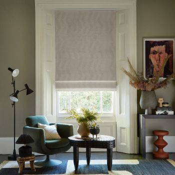 Pale beige fabric Roman blind lowered over a sash window, softly filtering daylight; cozy living room setting with green swivel armchair, textured coffee table, plants, muted walls and framed portrait.