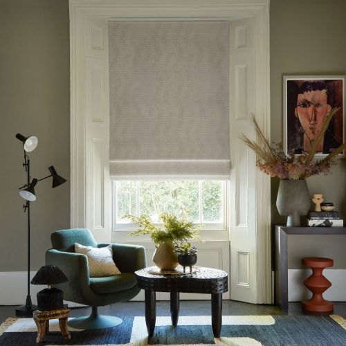 Pale beige fabric Roman blind lowered over a sash window, softly filtering daylight; cozy living room setting with green swivel armchair, textured coffee table, plants, muted walls and framed portrait.